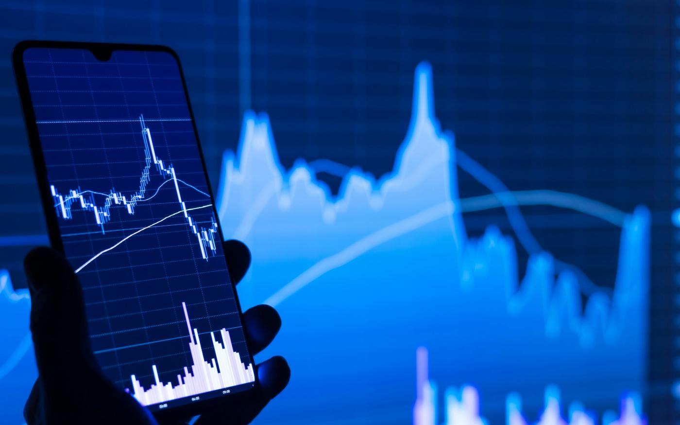 a person holding a cell phone in front of a stock chart by Adam Śmigielski courtesy of Unsplash.