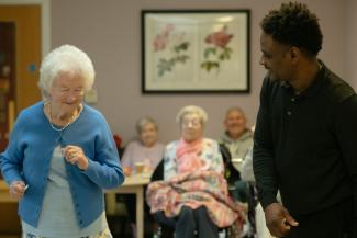 Elderly woman dances with caregiver in a community room by Age Cymru courtesy of Unsplash.